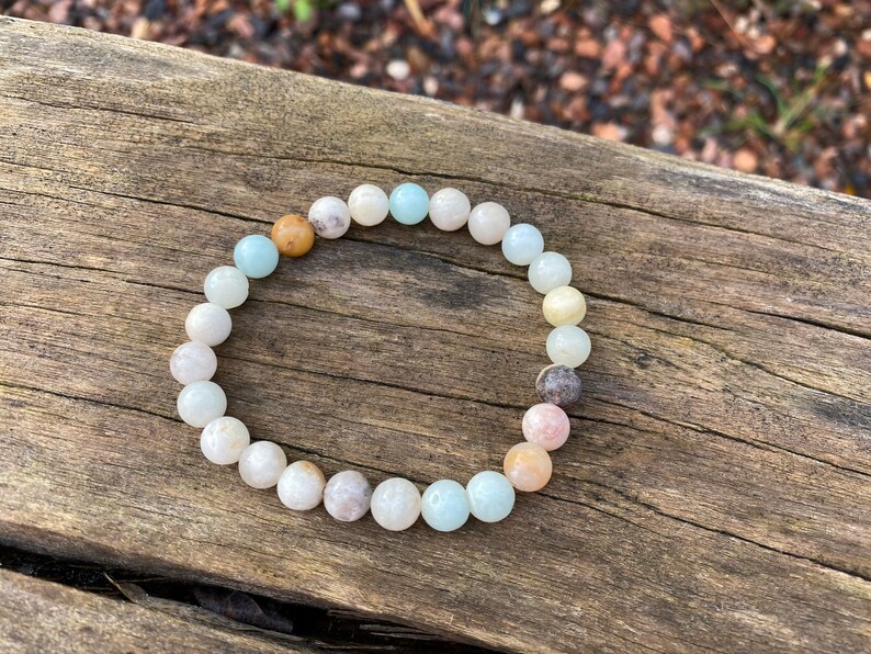 Puede incluir: Un brazalete hecho de cuentas de color azul claro, blanco y rosa. Las cuentas son redondas y tienen una forma natural ligeramente irregular. El brazalete est&aacute; sobre una superficie de madera.