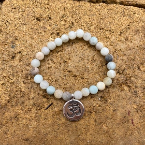 Puede incluir: Un brazalete de cuentas azul claro y blanco con un dije de Om plateado. El brazalete est&aacute; sobre un fondo de piedra marr&oacute;n.