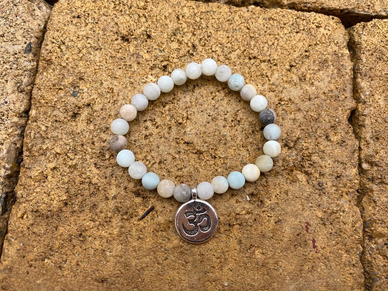 Puede incluir: Un brazalete de cuentas azul claro y blanco con un dije de Om plateado. El brazalete est&aacute; sobre un fondo de piedra marr&oacute;n.