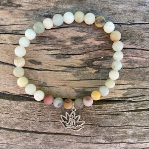 Puede incluir: Un brazalete de cuentas blancas y marrones con un dije de plata de una persona sentada en posici&oacute;n de loto. El brazalete est&aacute; sobre una superficie de madera.