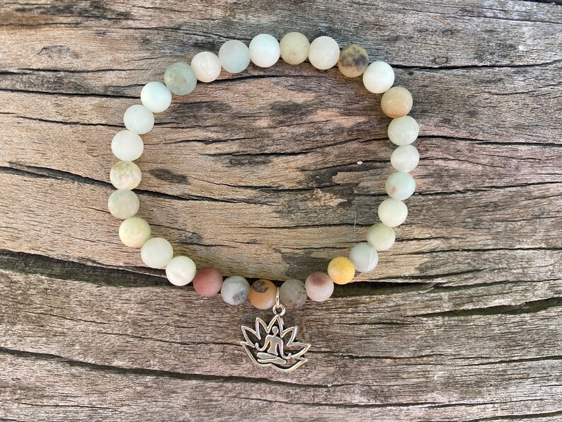 Puede incluir: Un brazalete de cuentas blancas y marrones con un dije de plata de una persona sentada en posici&oacute;n de loto. El brazalete est&aacute; sobre una superficie de madera.