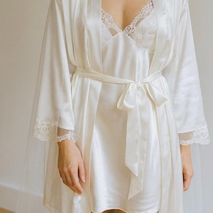 May include: Ivory satin robe and slip dress set. The robe has long sleeves with lace trim at the cuffs and a matching tie belt. The slip dress features a lace-trimmed neckline. A sheer veil is visible.