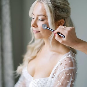 Puede incluir: Una maquilladora aplica colorete en la mejilla de una persona con un pincel. La persona tiene el pelo rubio y lleva un vestido blanco con adornos de perlas. La escena está iluminada con luz natural suave.