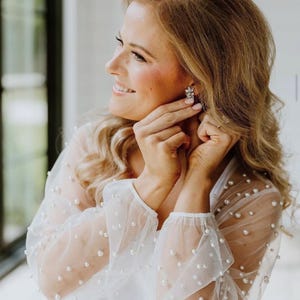 May include: A woman wearing a white sheer top with pearl embellishments on the sleeves. She is wearing a pair of small, sparkly earrings. Her blonde hair is styled in loose waves.