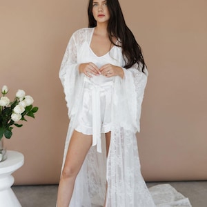 Op de afbeelding: Een witte satijnen pyjamaset met een bijpassende kanten kimono. De kimono heeft lange mouwen en een vloeiende sleep. Het model draagt sandalen met parels. De achtergrond is een neutraal beige.