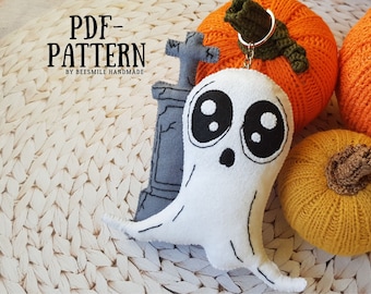 May include: A white ghost plush toy with black eyes and a mouth, attached to a key ring. The ghost is sitting on a woven surface with two knitted pumpkins in the background.