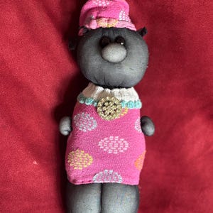 Puede incluir: Muñeca hecha a mano con cabeza y cuerpo negros, vestida con un vestido y sombrero rosas con estampado floral. La muñeca tiene un cuello blanco con una broche decorativa. La muñeca tiene ojos negros de botón y nariz redonda.