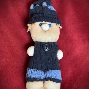 Puede incluir: Muñeca hecha a mano con cuerpo de tela, vestida con un conjunto de punto azul marino con rayas azul claro y un gorro a juego. La muñeca tiene un botón en la parte delantera, nariz blanca, ojos azules y pelo de tela color canela.