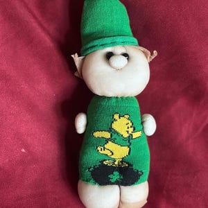 Może przedstawiać: Ręcznie robiona lalka z zieloną dzianinową czapką i swetrem. Lalka ma białą twarz, małe czarne oczy i brązowe uszy. Zielony sweter ma żółtą grafikę przedstawiającą misia i garnek złota. Ciało i stopy lalki są białe.