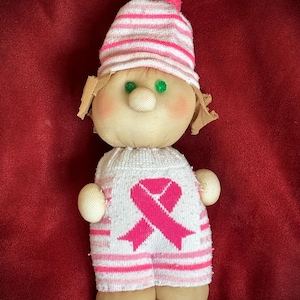 Puede incluir: Muñeca hecha a mano con un diseño de lazo rosa, que lleva un sombrero y un atuendo a rayas. La muñeca tiene ojos verdes, nariz beige y mejillas rosadas. El atuendo de la muñeca tiene un lazo rosa, que simboliza la concienciación sobre el cáncer de mama.