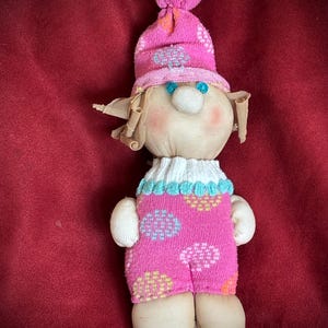 Puede incluir: Muñeca hecha a mano con sombrero y atuendo rosa, con cuello blanco y ojos azules. El atuendo de la muñeca tiene un estampado floral en varios colores. La muñeca está sobre un fondo rojo.