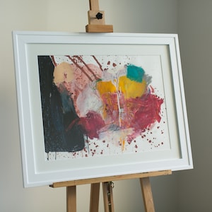 May include: Abstract painting in a white frame on an easel. The painting features splashes of red, yellow, blue, and white paint on a white background.