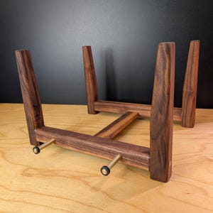 May include: A dark brown wooden stand featuring four tapered legs connected by a central crossbar. The front section includes two horizontal metal rods with small black rubber caps at the ends, designed to support an object in an upright position.