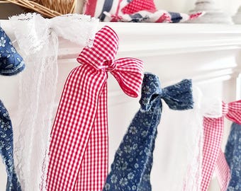 Puede incluir: Una guirnalda patriótica con lazos de cuadros rojos y blancos, encaje blanco y tela floral azul. Una estrella roja y una cesta están en el fondo. La guirnalda se exhibe en una repisa blanca.