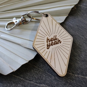 May include: A wooden diamond-shaped keychain with a sunburst design and the text "hell yeah" in a retro font.