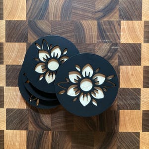 May include: A stack of four black coasters with a laser-cut floral design. The coasters have a circular shape with a light-colored wood grain visible through the cut-out flower pattern. The coasters are on a wooden cutting board.