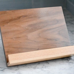May include: A wooden cookbook stand with a walnut-coloured base and a lighter wood shelf. The stand is angled to hold a book open.