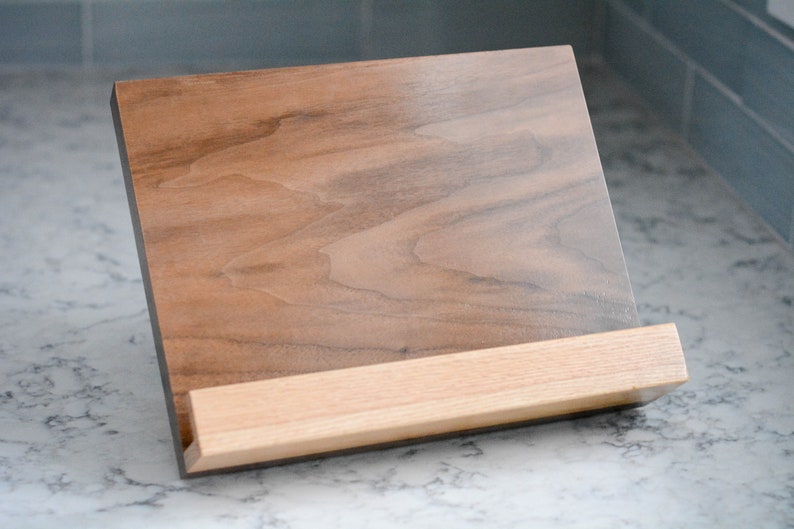 May include: A wooden cookbook stand with a walnut-coloured base and a lighter wood shelf. The stand is angled to hold a book open.