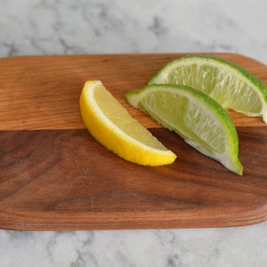 May include: A wooden cutting board with a light brown and dark brown wood grain pattern. There are three lime wedges and one lemon wedge on the board.