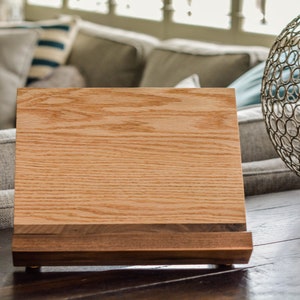 May include: A wooden phone stand with a natural wood finish. The stand has a raised platform for holding a phone or tablet and a lower shelf for storing items. The stand is made of two different types of wood, with a darker wood trim around the edges.