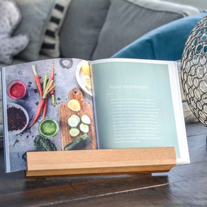 May include: A wooden book stand holding a cookbook open to a page titled "liquid nourishment". The page features a photo of fresh vegetables and fruit, including carrots, cucumbers, and berries.