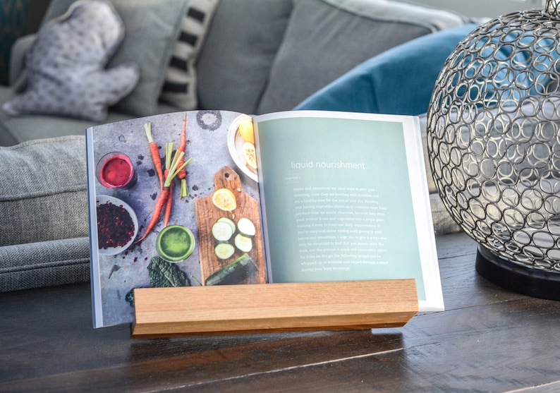 May include: A wooden book stand holding a cookbook open to a page titled "liquid nourishment". The page features a photo of fresh vegetables and fruit, including carrots, cucumbers, and berries.