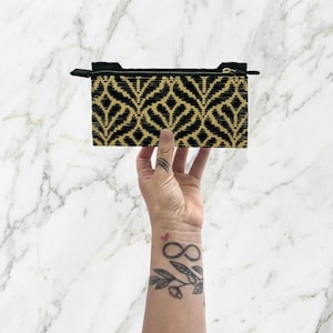 Peut inclure: Une pochette rectangulaire à fermeture éclair avec un motif géométrique répétitif en éventail noir et doré. Le tissu présente un tissage texturé et le bord supérieur est doté d'une fermeture éclair noire sur toute la largeur de l'objet.