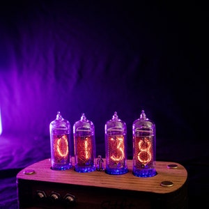 May include: A wooden clock with four glowing orange Nixie tubes displaying the time 01:38. The clock has a dark wood base with two knobs and the text "Clock Works" inscribed on the front.