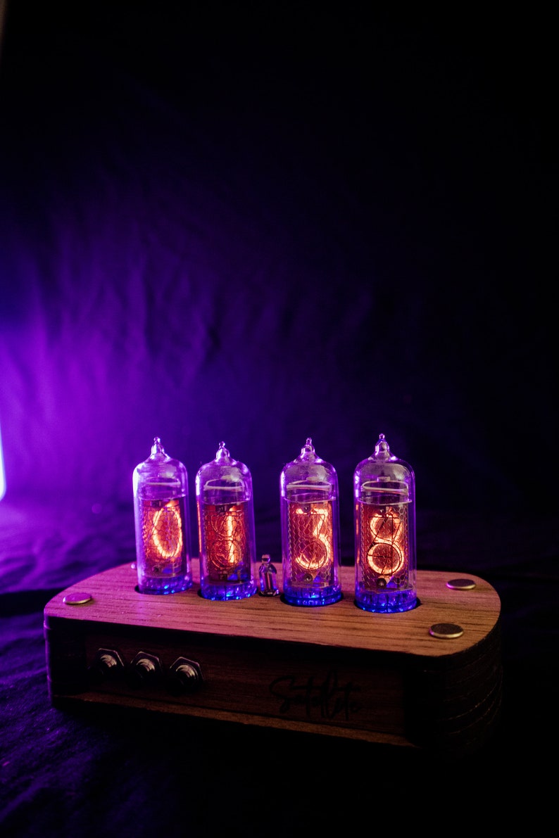 May include: A wooden clock with four glowing orange Nixie tubes displaying the time 01:38. The clock has a dark wood base with two knobs and the text "Clock Works" inscribed on the front.