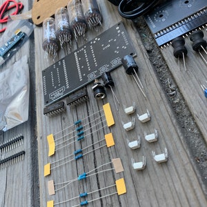 May include: A kit for building a clock using vacuum tubes. The kit includes a circuit board, vacuum tubes, resistors, switches, and a power supply. The circuit board is black with white text and markings. The vacuum tubes are glass with metal pins. The resistors are blue and brown. The switches are white. The power supply is black with a power cord.
