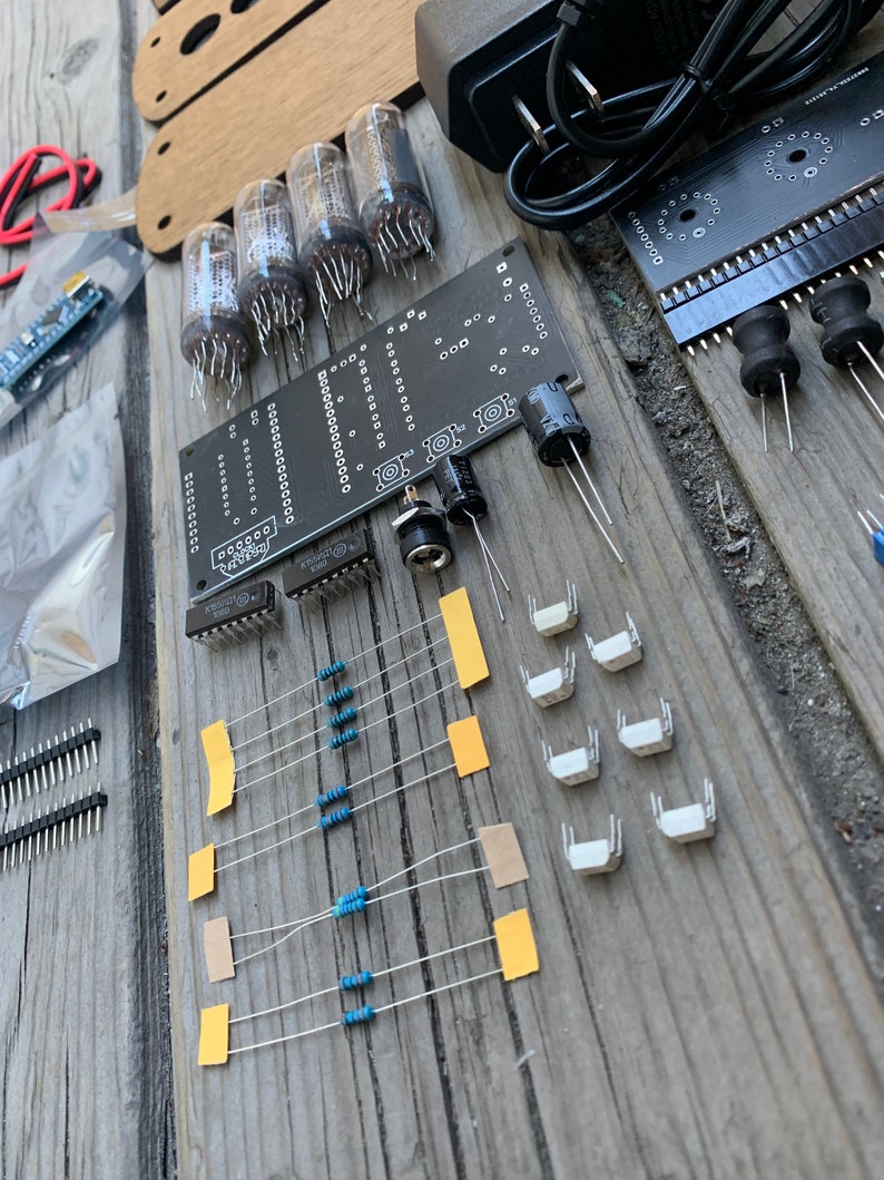 May include: A kit for building a clock using vacuum tubes. The kit includes a circuit board, vacuum tubes, resistors, switches, and a power supply. The circuit board is black with white text and markings. The vacuum tubes are glass with metal pins. The resistors are blue and brown. The switches are white. The power supply is black with a power cord.
