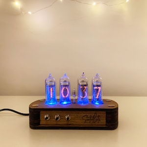 May include: A wooden clock with four Nixie tubes displaying the time 10:37. The clock has a dark wood finish and a copper accent on the front. The clock is lit with a blue glow.