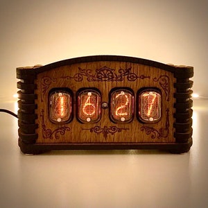 May include: A wooden digital clock with four glowing orange Nixie tubes displaying the time 06:27. The clock has a decorative wooden frame with ornate carvings and a dark brown border. The clock is powered by a black cord.