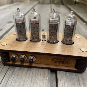 May include: A wooden clock with four clear glass Nixie tubes displaying the time. The clock has three black buttons and the text "Satellite" is engraved on the front.