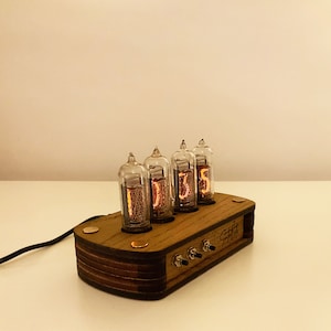 May include: A wooden clock with four clear glass Nixie tubes displaying the time "01:35". The clock has a brown wooden base with a label that reads "Sixty Five" and five toggle switches.