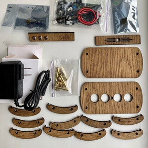 May include: A wooden kit for building a custom game console. The kit includes a variety of wooden pieces, including a top and bottom panel, buttons, and a control panel. The kit also includes a small bag of gold-colored screws and a small circuit board.