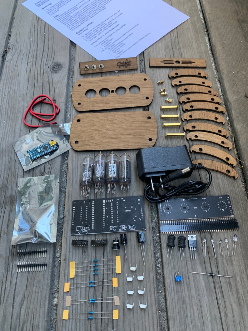 May include: A kit for building a Nixie tube clock. The kit includes a black circuit board, four Nixie tubes, a power supply, a set of wooden panels, and various electronic components. The kit also includes a set of instructions.