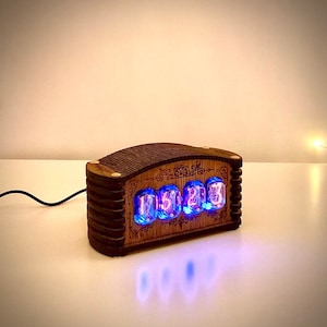 May include: A wooden digital clock with four glowing blue Nixie tubes displaying numbers. The clock has a dark brown, rectangular wooden frame with decorative carvings. A black power cord extends from the back.