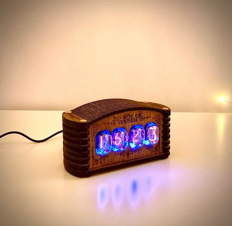 May include: A wooden digital clock with four glowing blue Nixie tubes displaying numbers. The clock has a dark brown, rectangular wooden frame with decorative carvings. A black power cord extends from the back.