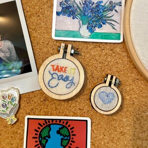 Könnte beinhalten: Eine Pinnwand mit verschiedenen Gegenständen, darunter Fotos, Aufkleber und kleine Stickrahmen. Einer der Rahmen trägt den Text "Take it Easy", ein anderer ein blaues Herz. Es gibt auch einen Aufkleber mit einem Keith Haring-Design.