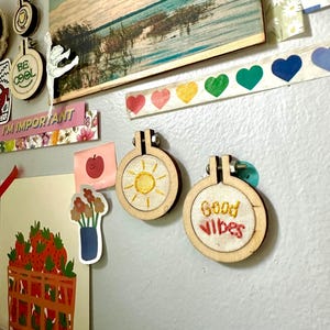 Op de afbeelding: Een verzameling decoratieve items op een grijze muur. Twee kleine houten borduurringen met geborduurde ontwerpen, een met een zon en de andere met de woorden "Good Vibes". Andere items zijn stickers, een landschapsafdruk en decoratieve tape.