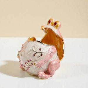 Pode incluir: Uma caixa de bugigangas decorativa em forma de sapo. O sapo é principalmente branco com detalhes em rosa e acabamento dourado. Possui olhos vermelhos em forma de joia e é adornado com pequenos cristais rosa e transparentes. O interior é de cor dourada quente.