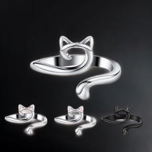 May include: A silver-colored cat ring with a wraparound design. The ring features a cat head and tail, with the tail wrapping around the finger. Additional rings in silver, silver with pink accents, and black are also shown.