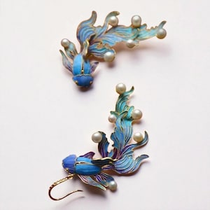 May include: A pair of goldfish earrings. The earrings are blue, purple, and gold, with small pearls. The fish design has flowing fins and tails. The earrings are an elegant accessory, suitable for special occasions.