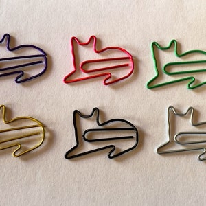 May include: Six colorful cat-shaped paper clips in purple, red, green, gold, black, and silver. The clips are arranged on a white surface, showcasing their playful design and vibrant hues. These clips are ideal for organizing documents.