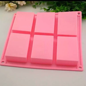 Könnte beinhalten: Eine rosa Silikon-Seifenform mit sechs rechteckigen Hohlräumen. Die Form hat eine einheitliche rosa Farbe, und die Hohlräume sind gleichmäßig verteilt. Die Form ist für die Herstellung von Seifenstücken oder ähnlichen Artikeln konzipiert.