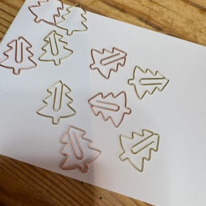 May include: A set of nine gold and rose gold metal Christmas tree paper clips. The paper clips are arranged on a white sheet of paper.
