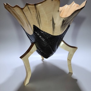 May include: A wooden bowl with a unique, organic shape and a black, textured base. The bowl is supported by three legs, also made of wood.