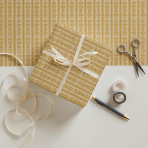 May include: A gift wrapped in yellow and white patterned paper with a white ribbon. The gift is on a white surface with a roll of tape, a pen, and scissors.