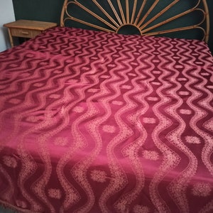 May include: A burgundy bedspread with a repeating wave pattern and floral accents. The bedspread is draped over a bed with a woven headboard and two matching nightstands. The fabric has a subtle sheen, suggesting a luxurious material.